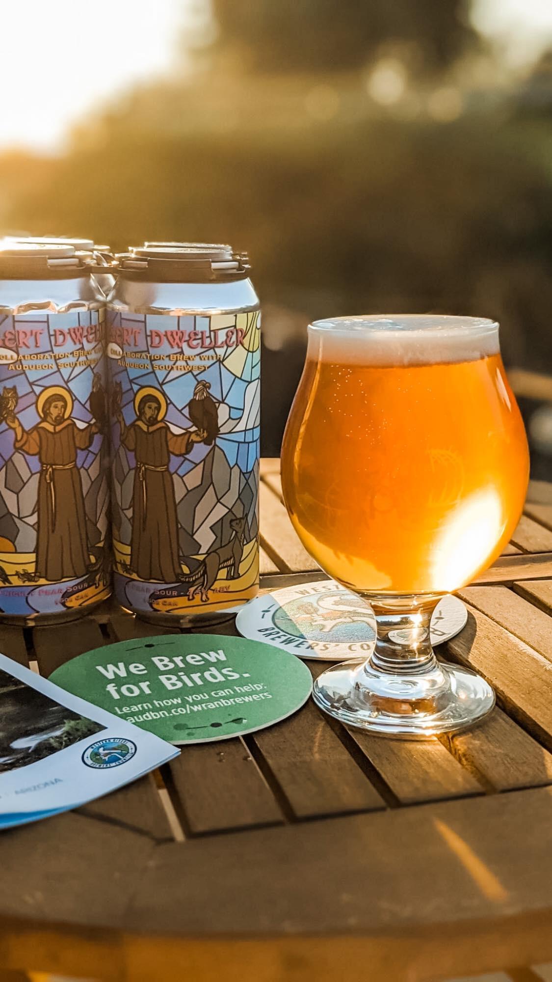 Cans of Desert Dweller Prickly Pear Sour behind a glass of the brew glowing in the sun and Western Rivers Brewers' Council coasters reading "we brew for birds".