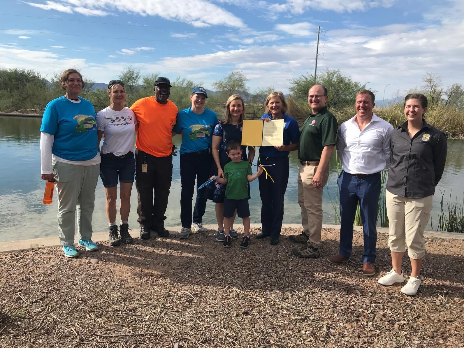 A group of people holding up a newly signed Urban Bird Treaty for the City of Phoenix stand in front of a still-watered wetland pond at the Rio Salado Habitat Restoration Area.
